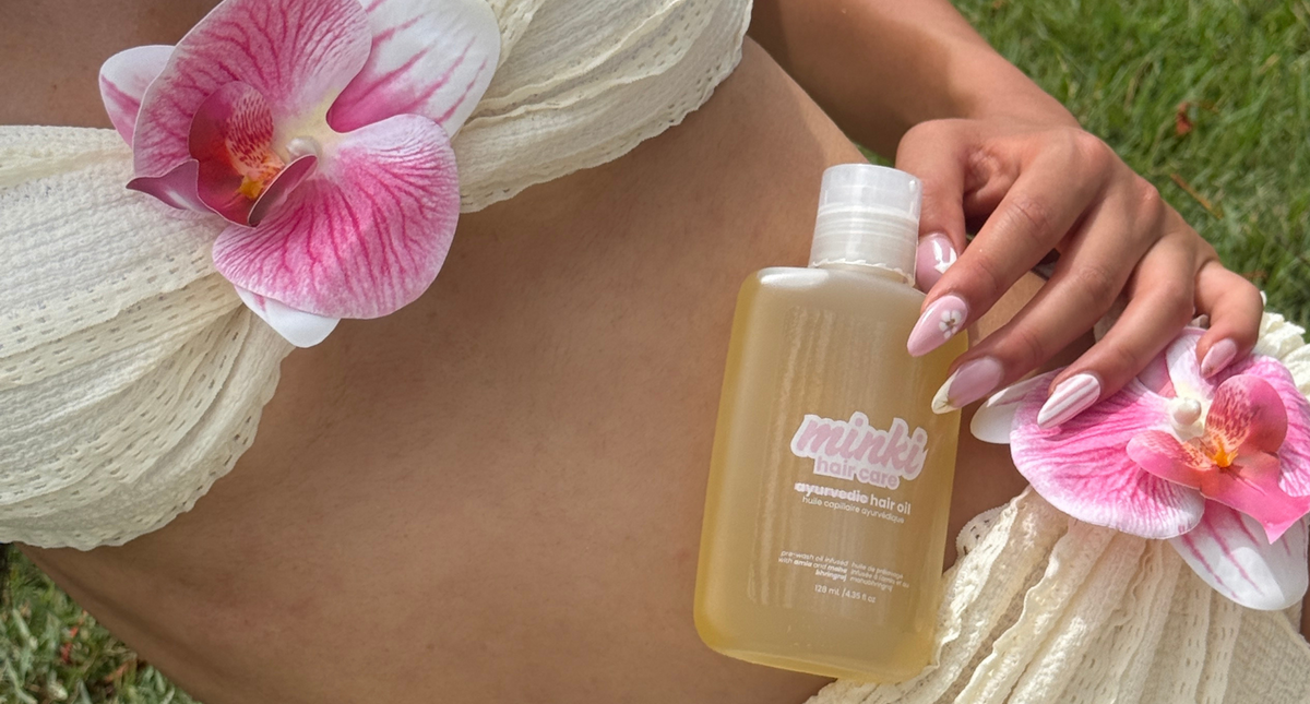 Ani, the founder, outdoors holding a bottle of Minki Hair Oil. The person is wearing a cream-colored orchid bikini, with green grass visible in the background.